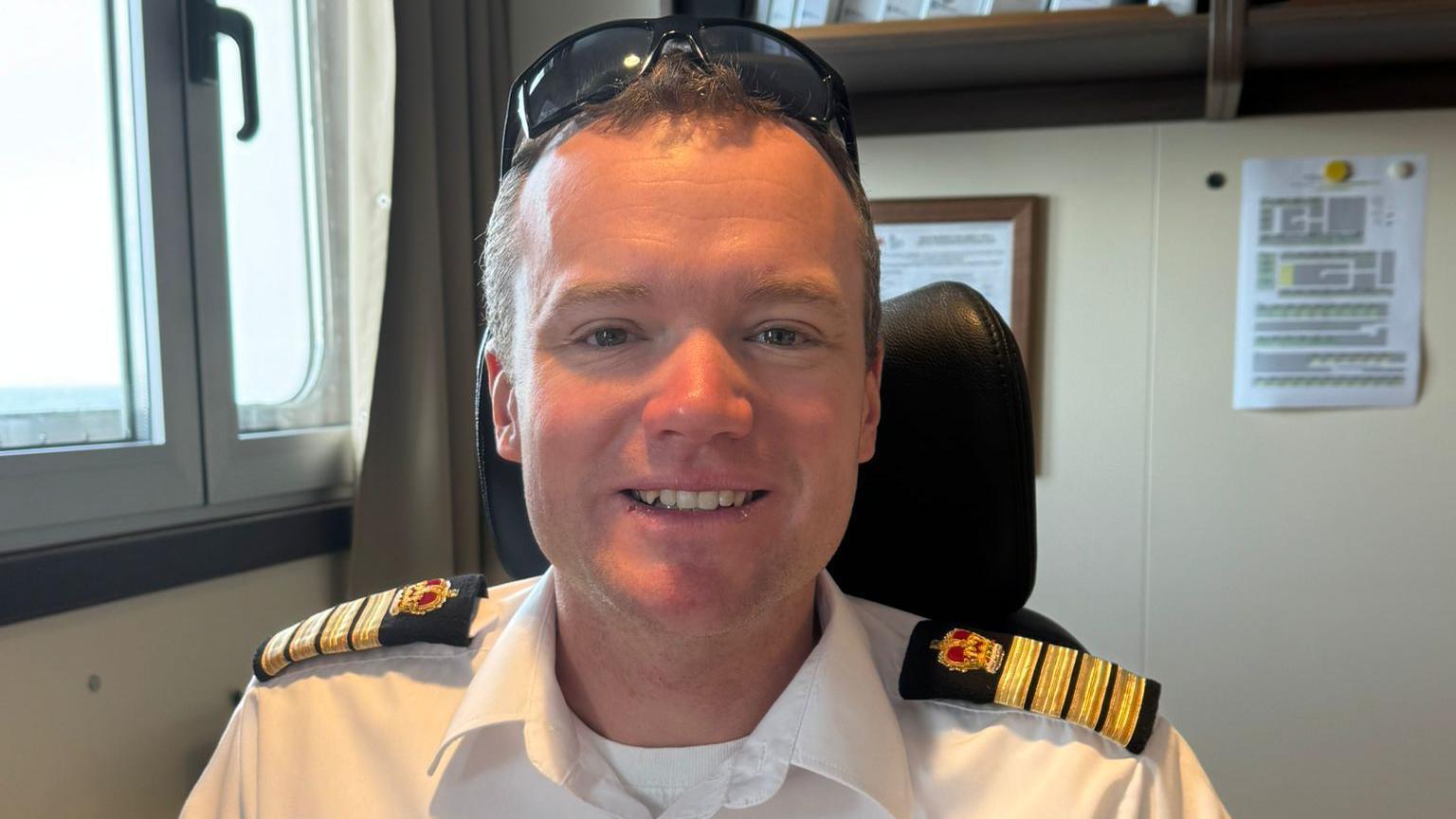 Capt Will Whatley is smiling at the camera. He has fair hair and sunglasses balanced on his head. He is wearing a white shirt with epaulettes. He is sitting in a chair on board a ship