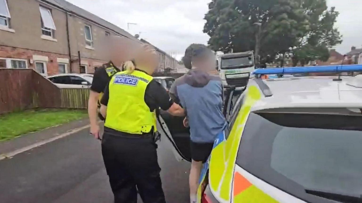 A man being put into the back of a police car. A female police officer is helping him inside. A second officer is standing next to her.