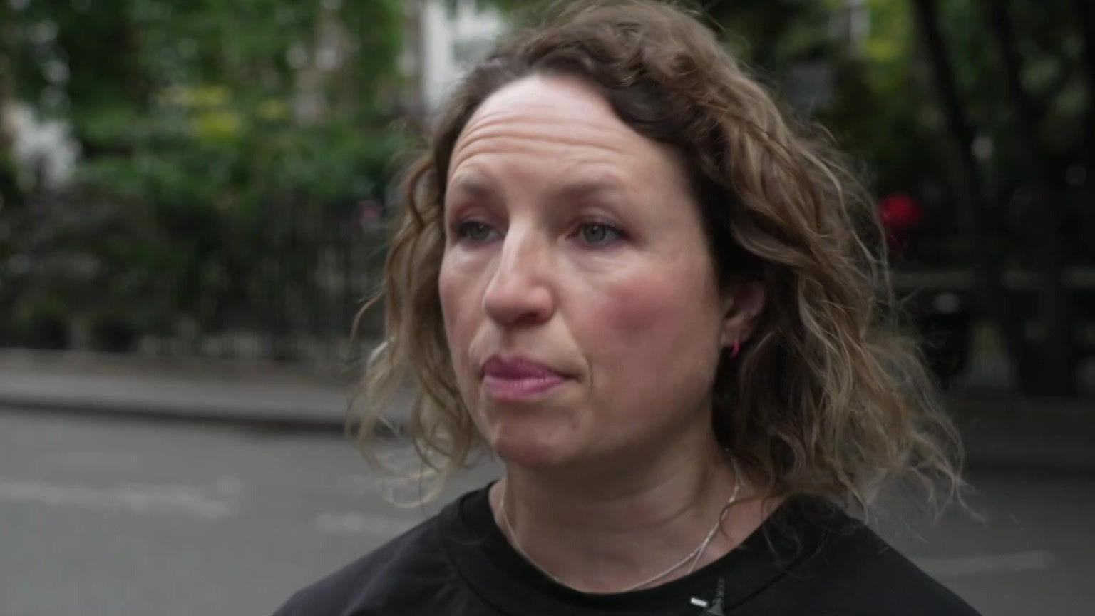 A woman with curly hair wearing a black top and microphone headset is speaking on a city street, with trees and railings in the background.