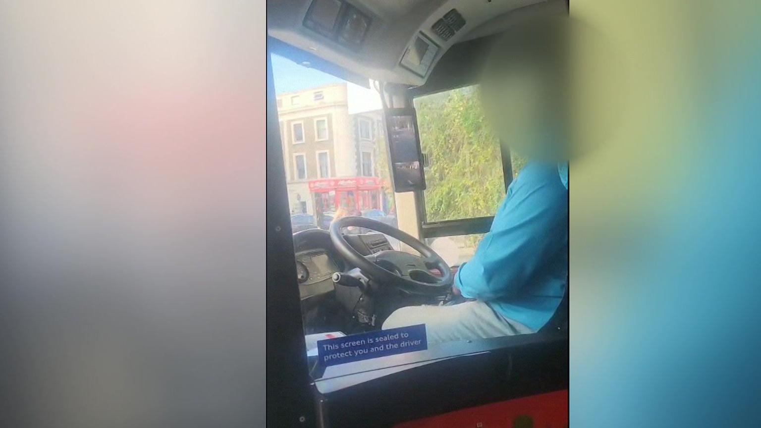 Mobile phone footage of a bus driver sitting in his cab. He is wearing light blue trousers and a blue shirt. His face has been blurred. 