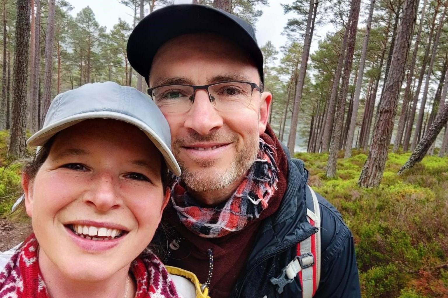 Ceris and Oliver Blomfield in a forest. They are both looking directly into the camera. Ceris is wearing a light blue baseball cap and Oliver has glasses, a navy cap and a navy jacket.