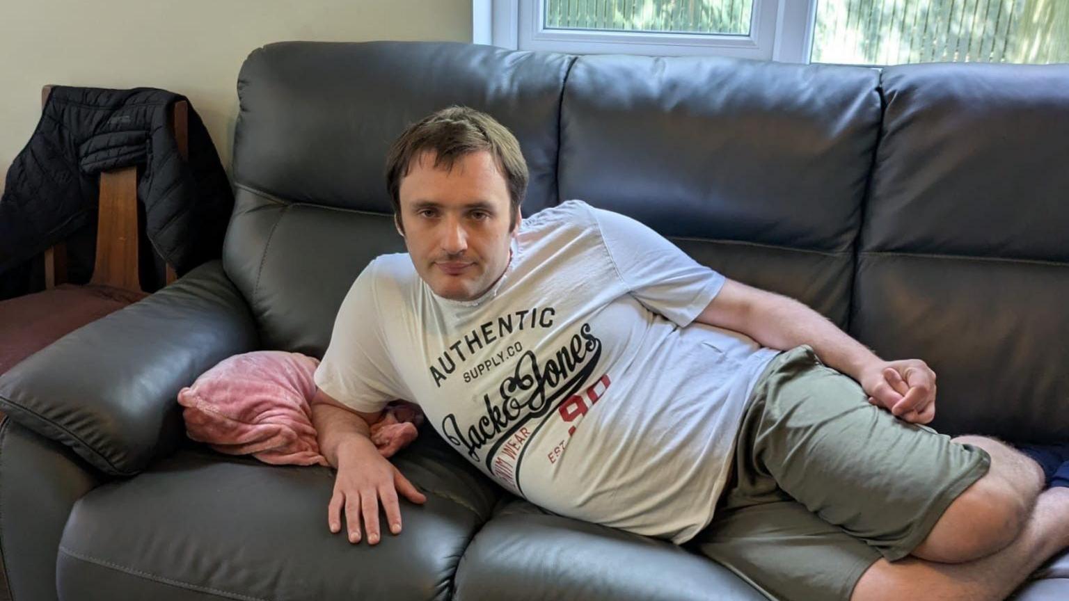 A man lies on a leather sofa. He is wearing green shorts and a white t-shirt. There is a pink cushion on the sofa. 
