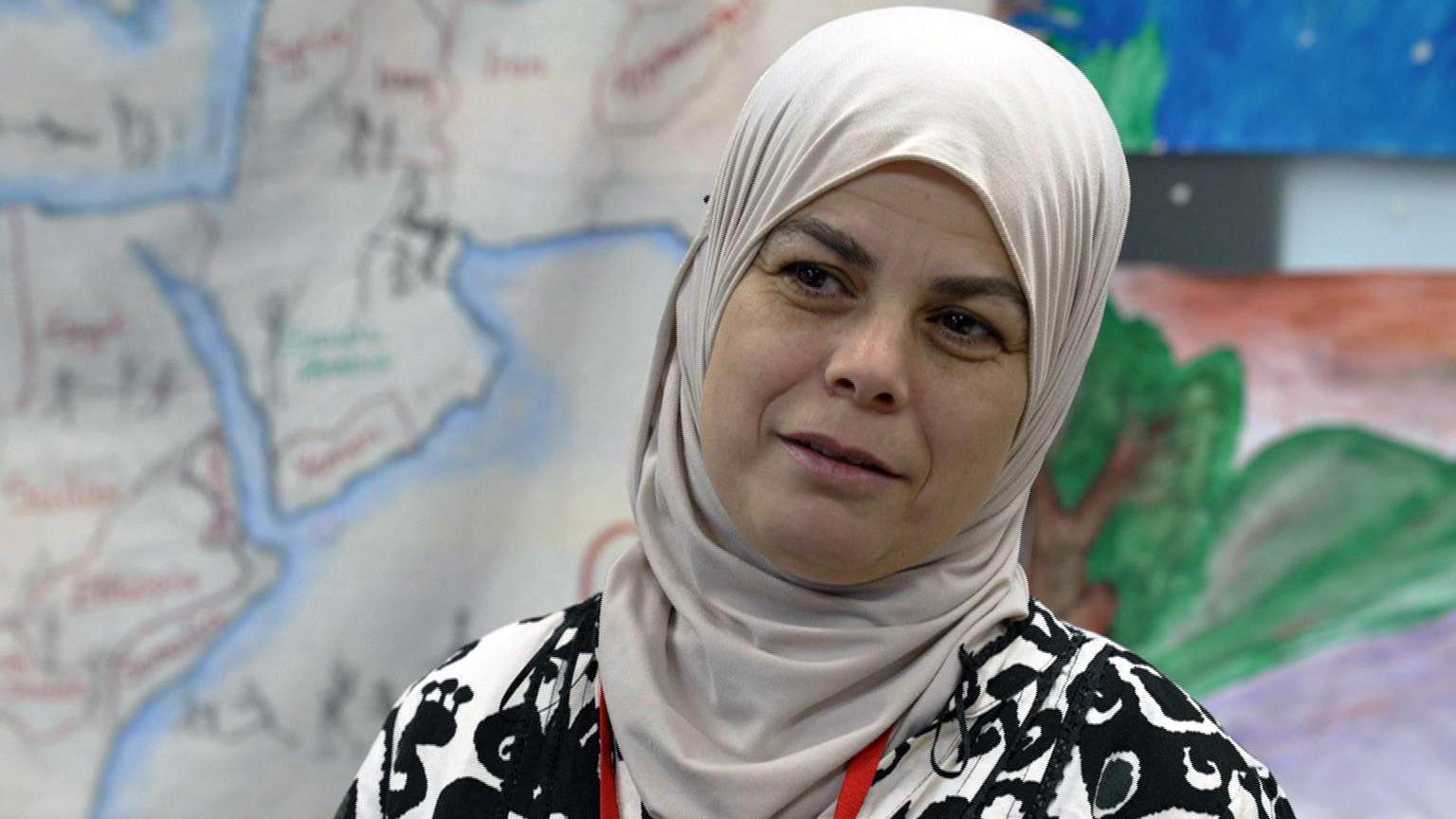 Warda is wearing a grey hijab around her head and neck and a black and white monochrome top. She is standing in front of a very large painting depicting a map.