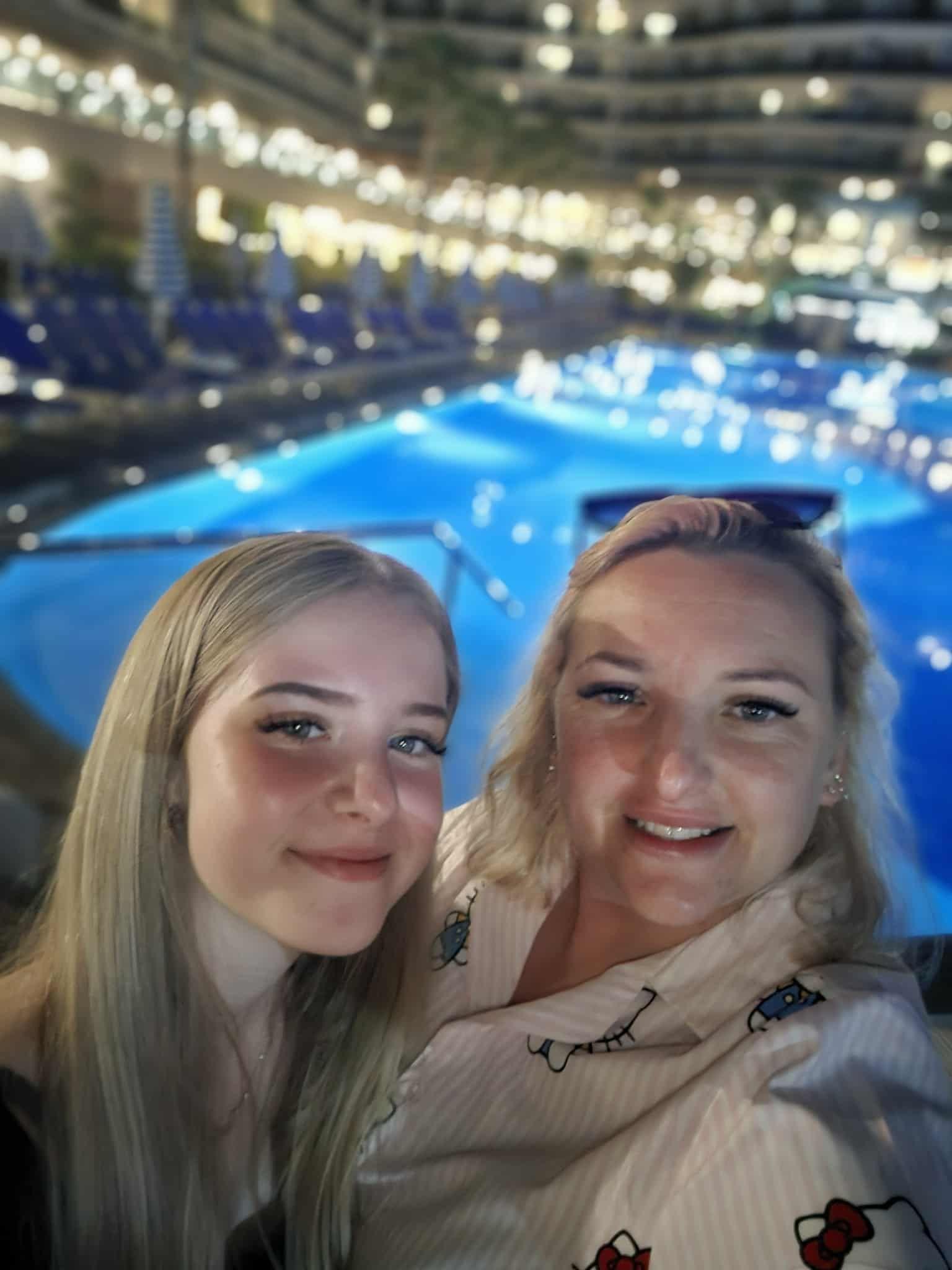 Lexi, left, and Becky right in a selfie in front of a large outdoor pool at night time with a hotel in the background. Lexi has long straight blonde hair and Becky has shoulder-length blonde hair.