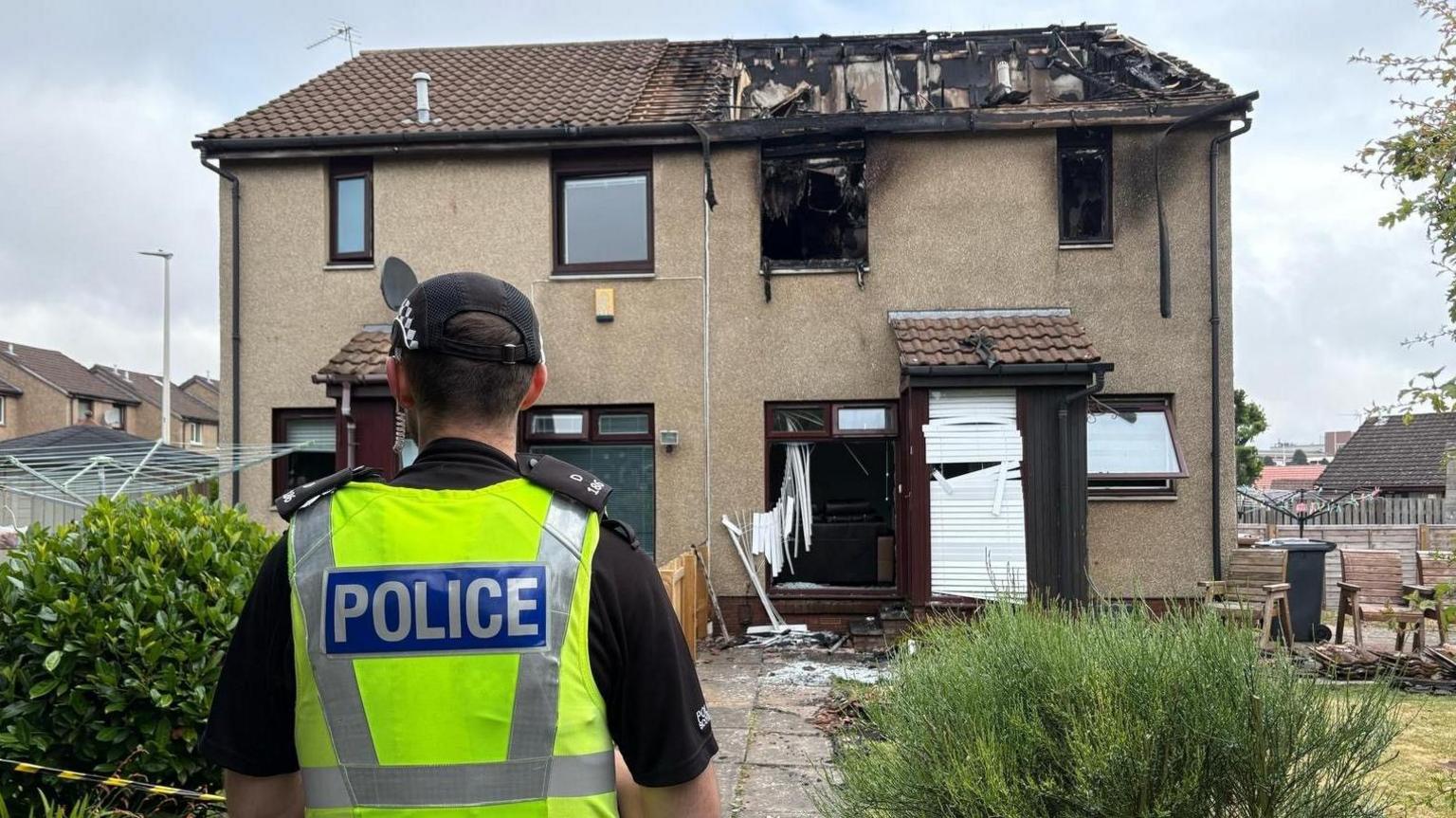 A police officer stand s with his back to the camera looking at a house which has been badly damaged by fire