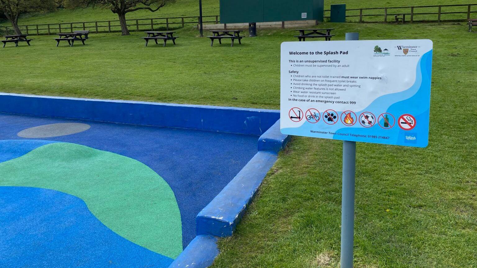A sign which says “Welcome to the Splash Pad” next to a grassy area and blue enclosed area