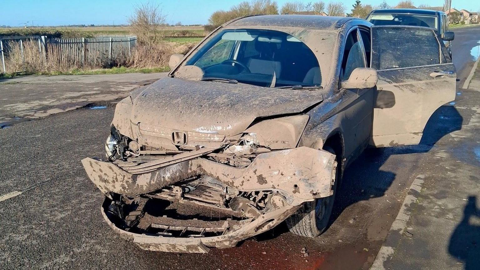 A dark-coloured Honda car caked in mud, its front bumper is hanging off and it has been parked by the side of a road in Cambridgeshire. The back left hand side door is wide open. Part of the windscreen has been wiped so it is not caked in brown dried mud. Fields can be seen in the background and a blue sky.