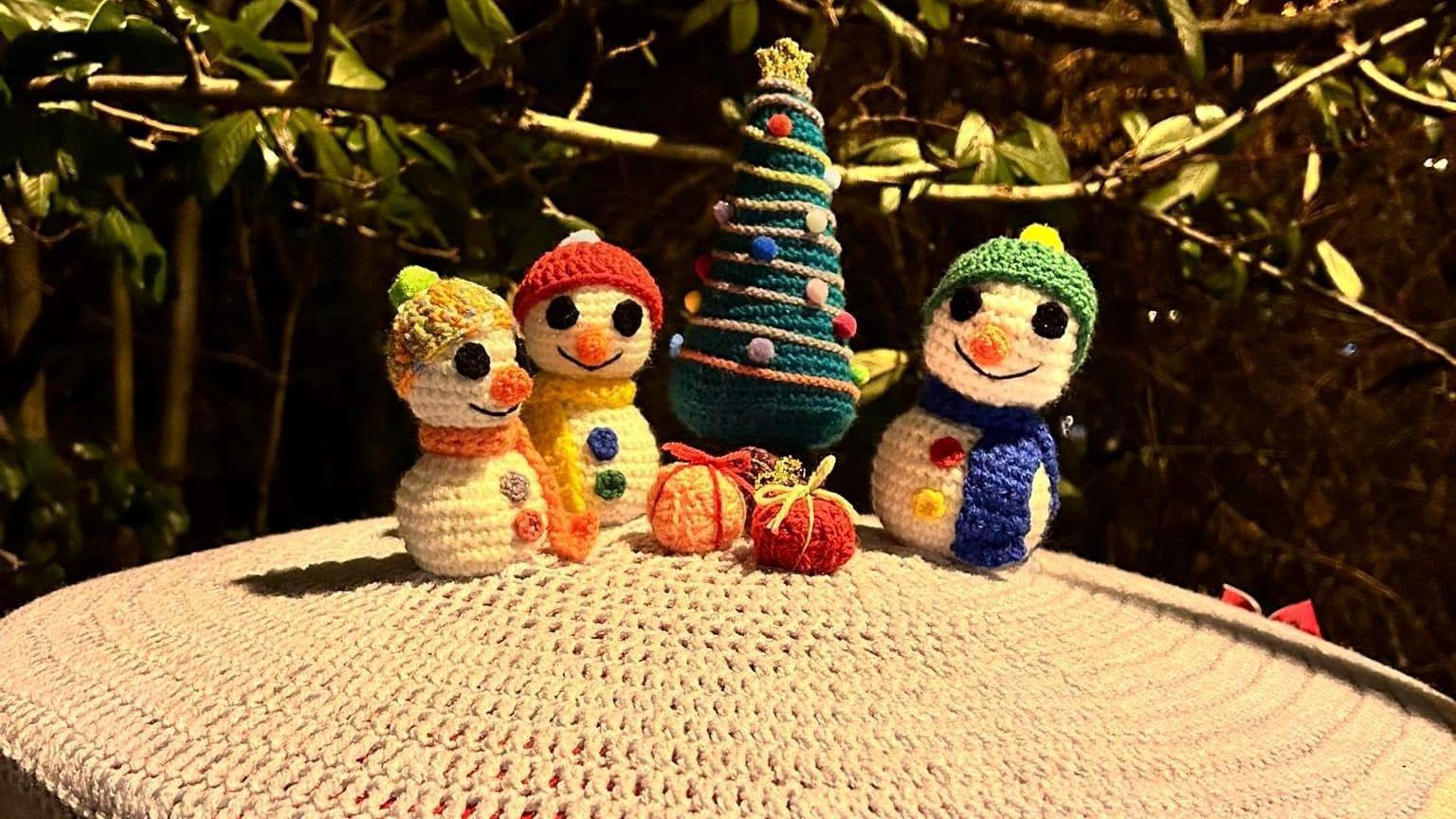 A white postbox topper is decorated with three snowmen in the middle, each one with different coloured buttons and scarves and a smiling face. There are presents on the ground, complete with ribbons, and a tree stands behind them with knitted tinsel and baubles.