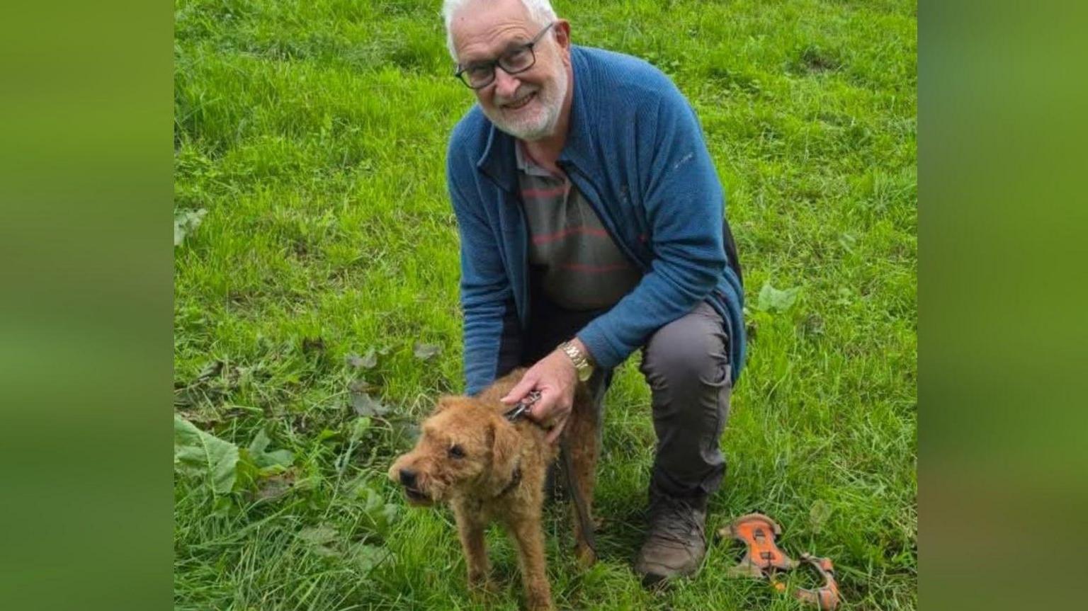 Mike McGrath crouched down and holding his Lakeland terrier Sherlock by his lead. They are in a grassy area and Mr McGrath is looking at the camera and smiling. He is wearing a blue fleece and grey trousers and has short white hair and a beard.