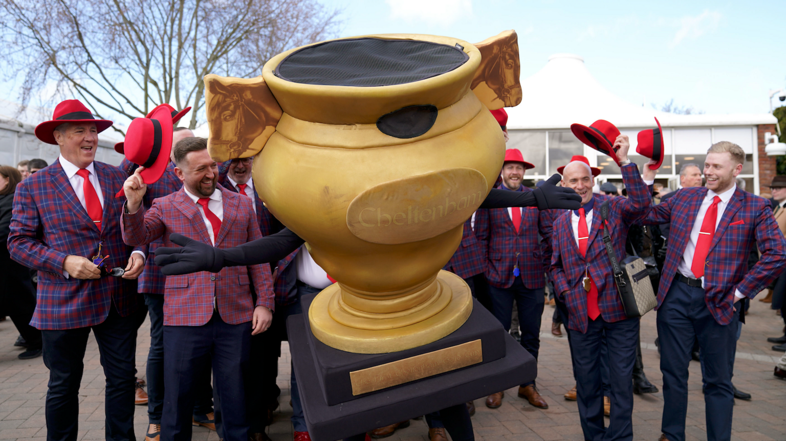 A group of men in red and blue checkered blazers with bright red hats. In the centre of the photo is a person dressed in a gold cup mascot suit. The men are holding their hats up and cheering with the mascot.