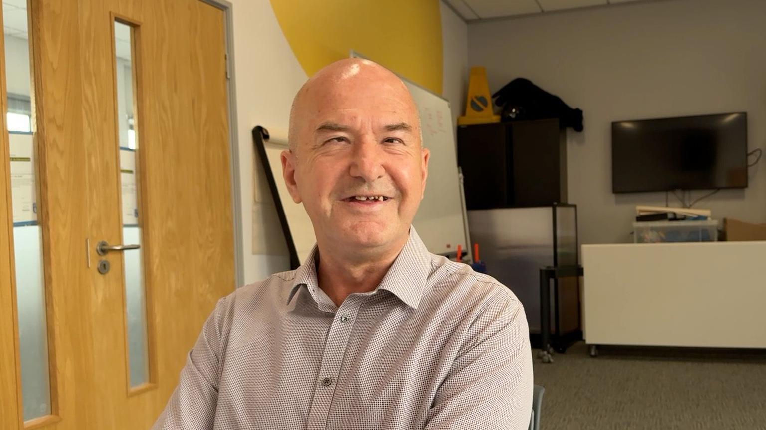Clive Wood is looking at the camera. He is wearing a grey shirt. 