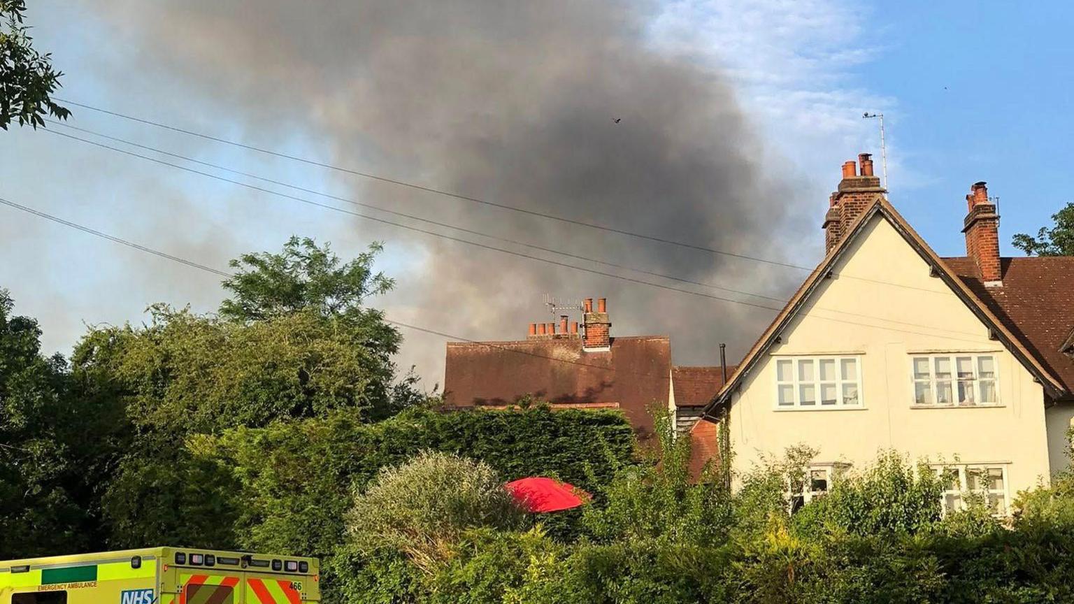 Black smoke from an industrial fire rising above houses and hedges in Letchworth Garden City. The roof of an ambulance can be seen on the right.