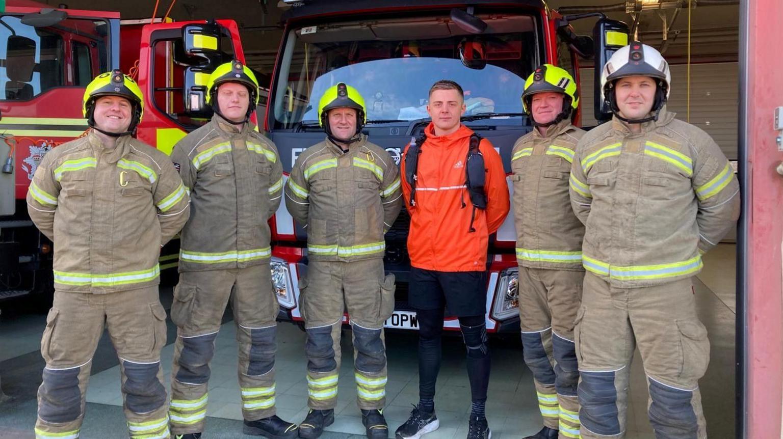 Mr Williams pictured in an orange jumper and black shorts and leggings. Around him are five firefights wearing their beige uniforms with yellow trim and hard hats. Behind them are two fire engines. 