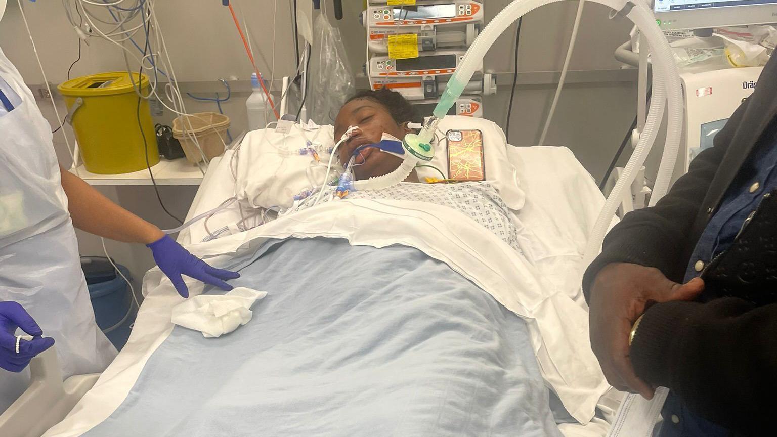 Ketia Moponda in her hospital bed. She is sleeping and has a huge tube coming out of her mouth. There are lots of wires surrounding her which are hooked up to a machine. We can see a nurse and a family member standing by her bed. But we only see there arms because they are cropped. 