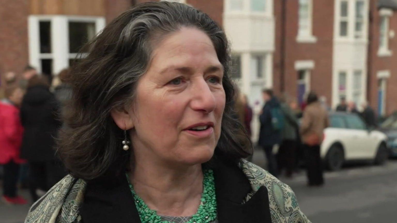Susan Walker is looking side on to the camera, she has dark hair and is wearing a coat with patterns, a green top and drop pearl earrings. In the background is a crowd of people who are out of focus. 
