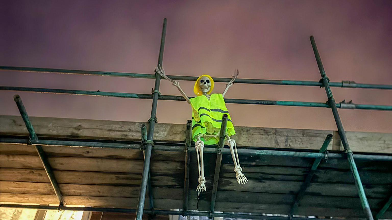 A skeleton in a hi vis jacket and a hard hat sitting on top of scaffolding. The night sky has a purple tinge and looks extra spooky.