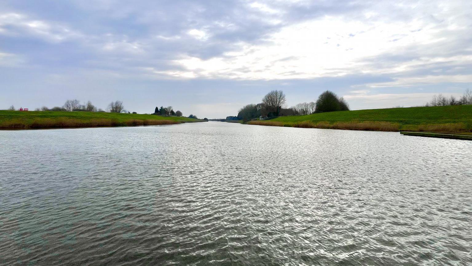 A body of water with green grassy banks either side, in the distance are a few trees, some of them sparse due to winter.