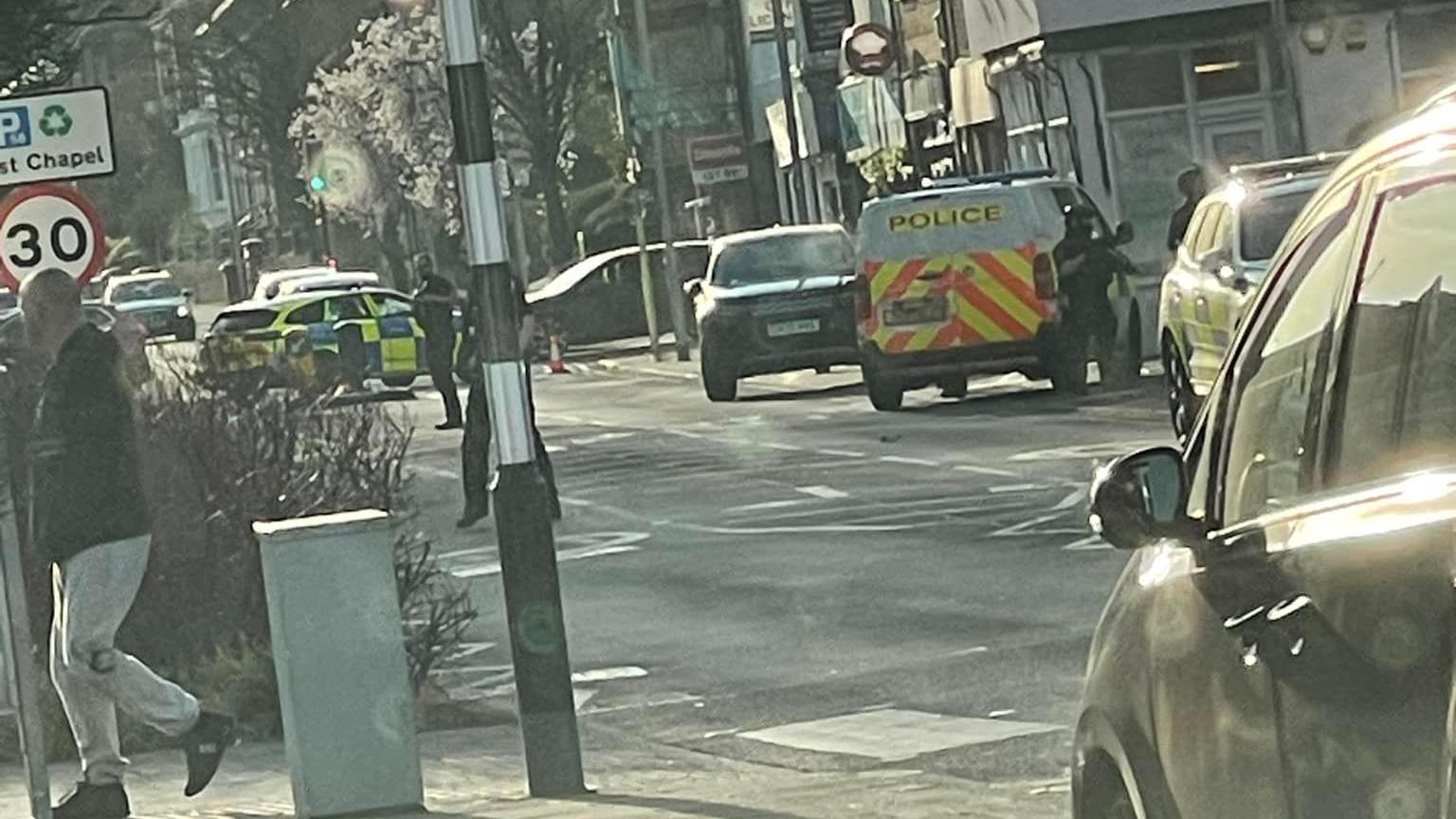 A photo taken from across the road shows three police vehicles, including a van, parked on the street. Armed police officers dressed in black vests and helmets can be seen