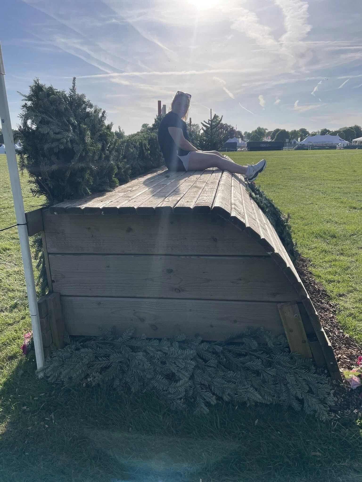 A young woman sits on a large wooden cross-country eventing obstacle