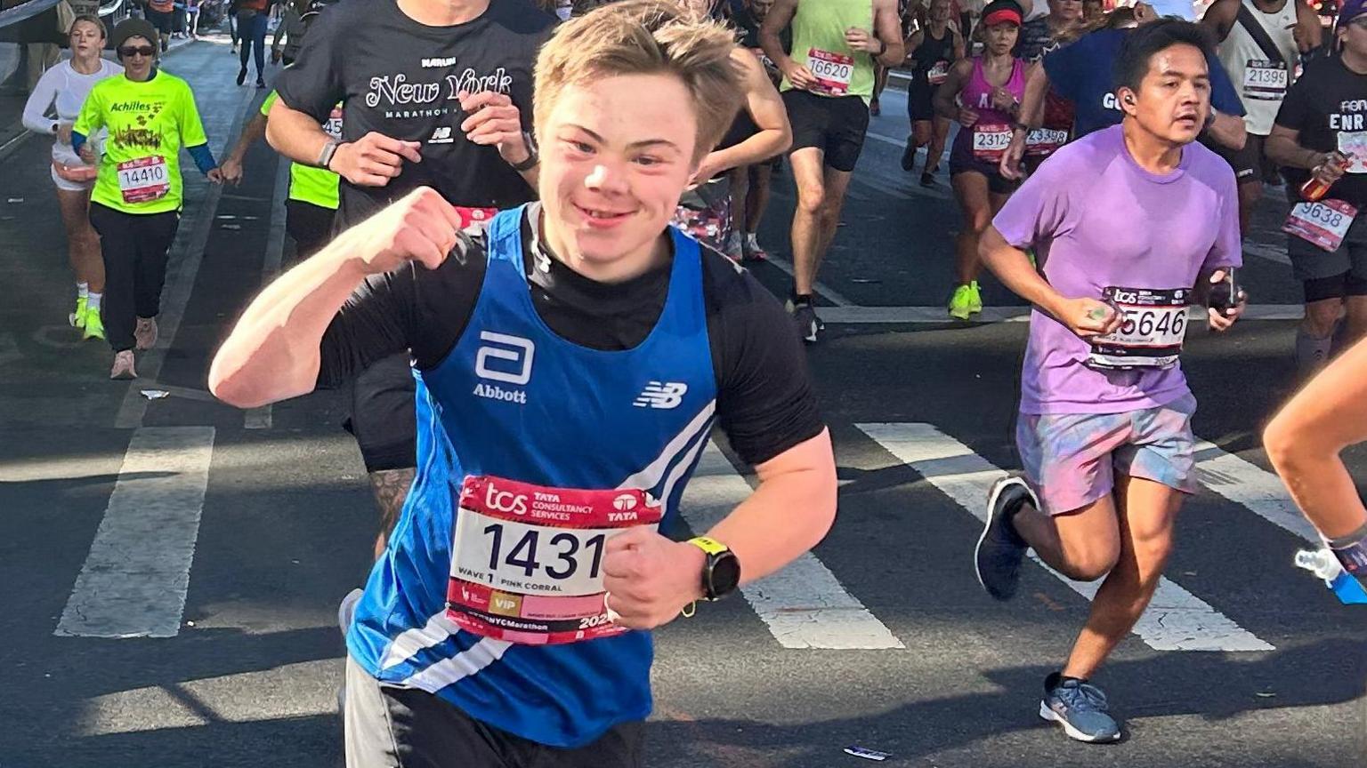 Lloyd Martin running the New York Marathon, posing for the camera with his fist in the air in a blue and black top. There are several runners behind him.