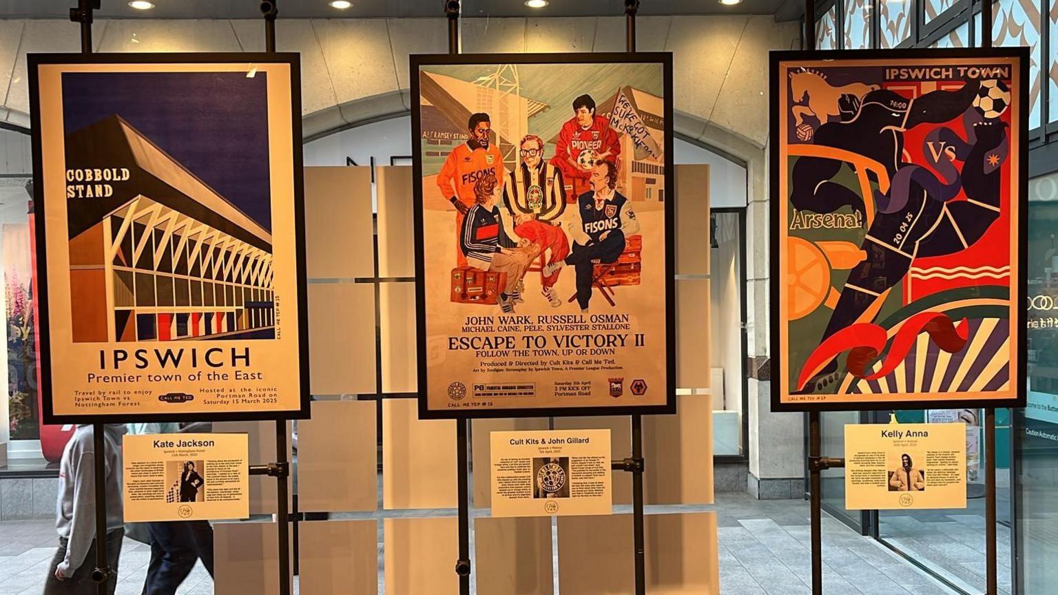 Three of the posters framed and on display in front of a long glass window. All of them have special art designs, all featuring something to do with Ipswich Town Football Club, including players, crests and the Portman Road stadium.