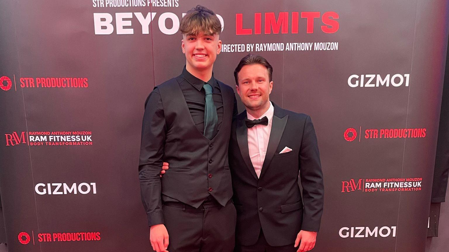 A tall young man wearing a black suit and a dark green tie and a slightly older man wearing a tuxedo. They're standing with their arms around each other and smiling at the camera, with a banner for their film behind them.