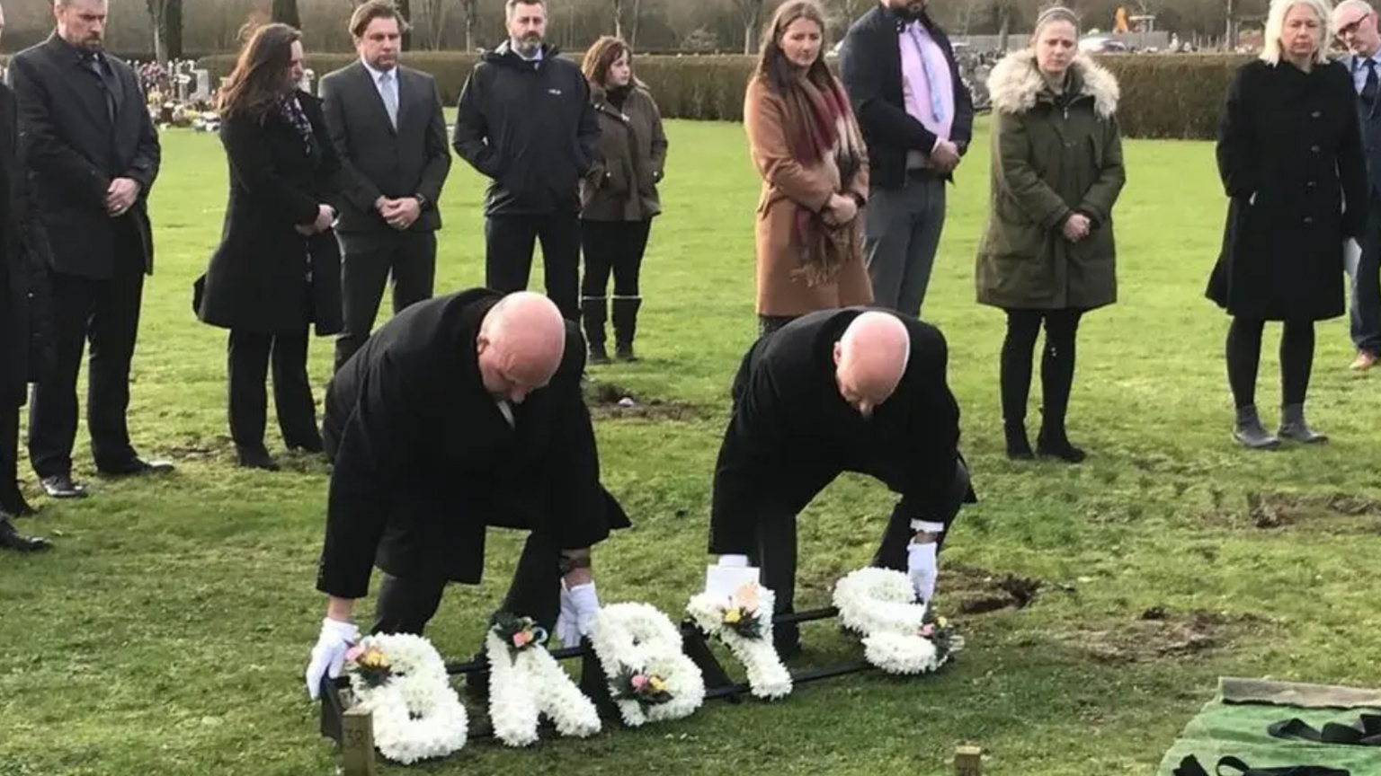 Mourners gather at a graveyard as two men lay a flower wreath which reads Baby S