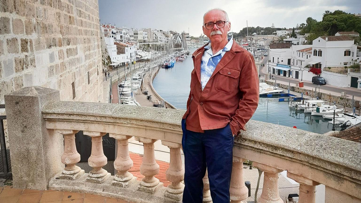 David is stood on the balcony of a stone building, overlooking a harbour where yachts and small boats are moored. There are rows of white buildings in the background. He is wearing a blue and white patterned shirt and a brown jacket and blue trousers, leaning against the balcony wall, with his hands in his pockets.