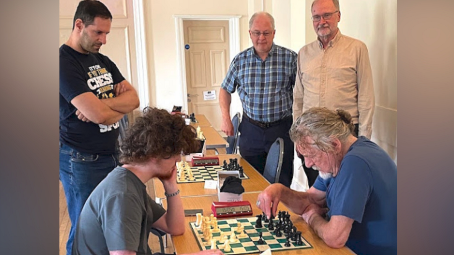 Someone wearing a grey T-shirt with curly hair is on the left playing chess with Plant, wearing a blue top on the right. Three other men are standing up - the one on the left with dark hair is wearing a blue top including words and the other two have grey hair.