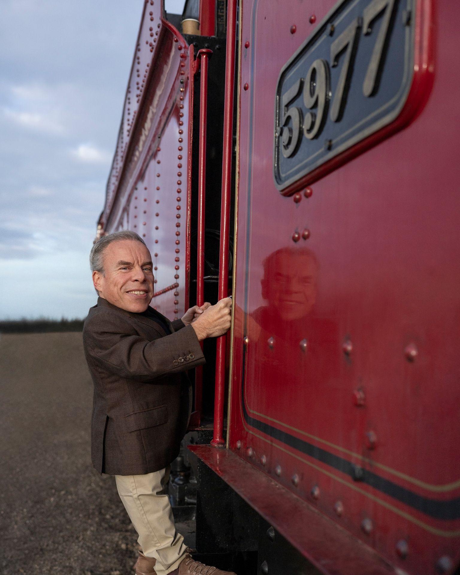 Warwick Davis standing on a step climbing into a steam train carriage