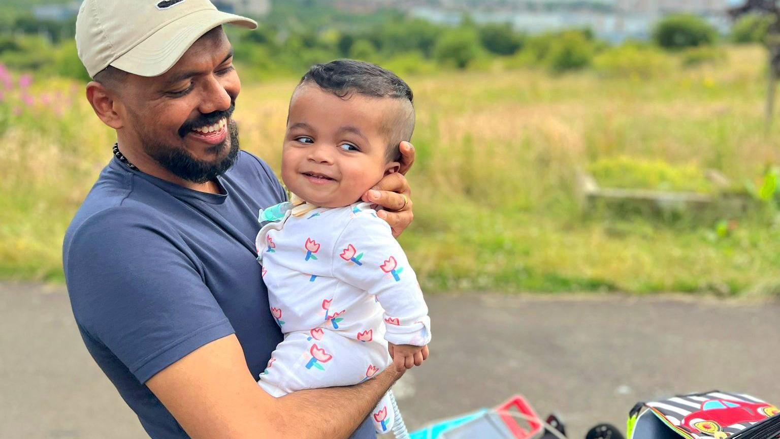 Gabriel's dad is wearing a blue t-shirt anf cap. He is holding his son in his arms and holding his head up. They are both smiling. There is green grass in the background.