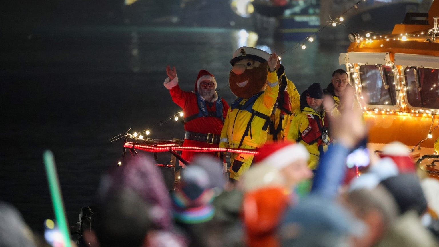 Father Christmas in his traditional red outfit stands next to a man wearing a lifeguard mascot costume, with yellow waterproofs and an oversized head with a brown beard on a lifeboat at night, while crowds of people wave from the shore.