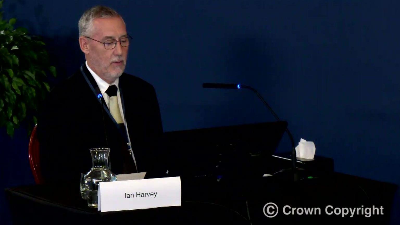 Ian Harvey, with short grey hair and light grey stubble, sits at a desk containing microphones and a computer screen