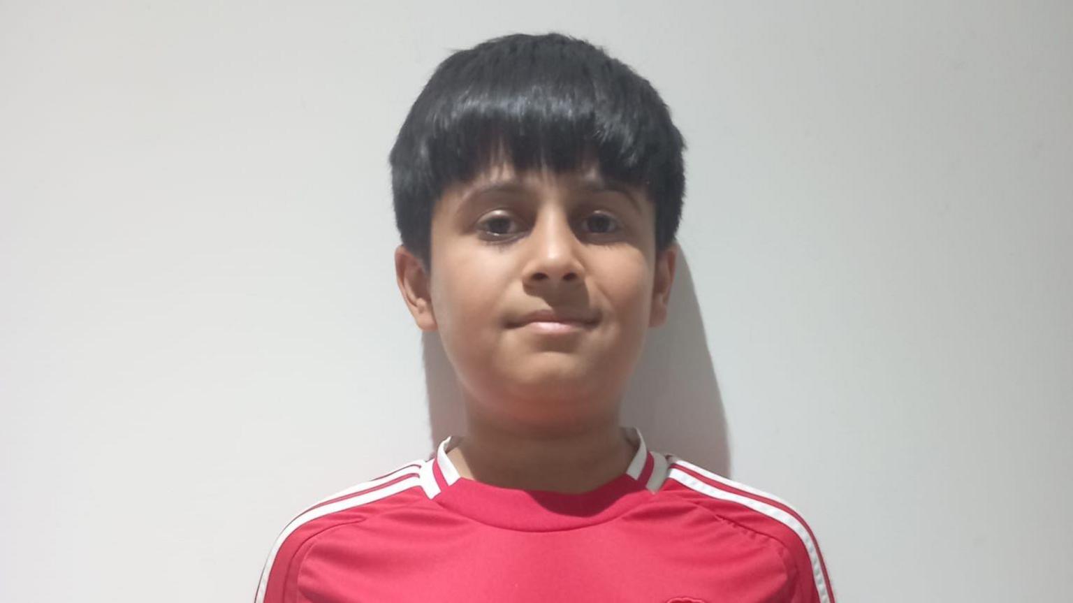 A boy with black hair in a Manchester United shirt standing against a white wall