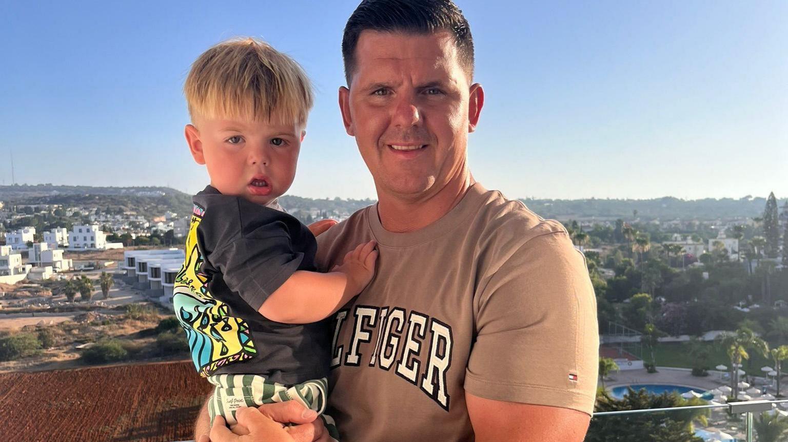 Teddie and his father, Ryan, pictured on holiday. Ryan is holding Teddie, Ryan is smiling at the camera and has a tanned t-shirt on with the words "Hilfiger" in block white writing across the front. He has dark cropped hair. His son has a charcoal t-shirt on with a colourful pattern on the back with striped green and white shorts. He has blonde hair. 