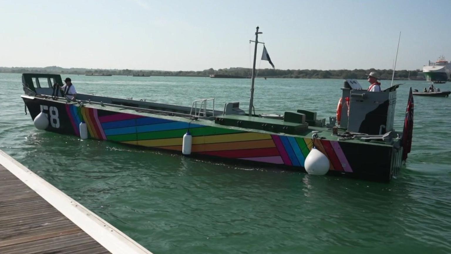 A landing craft approaching a jetty, with rainbow pride colours on its sides