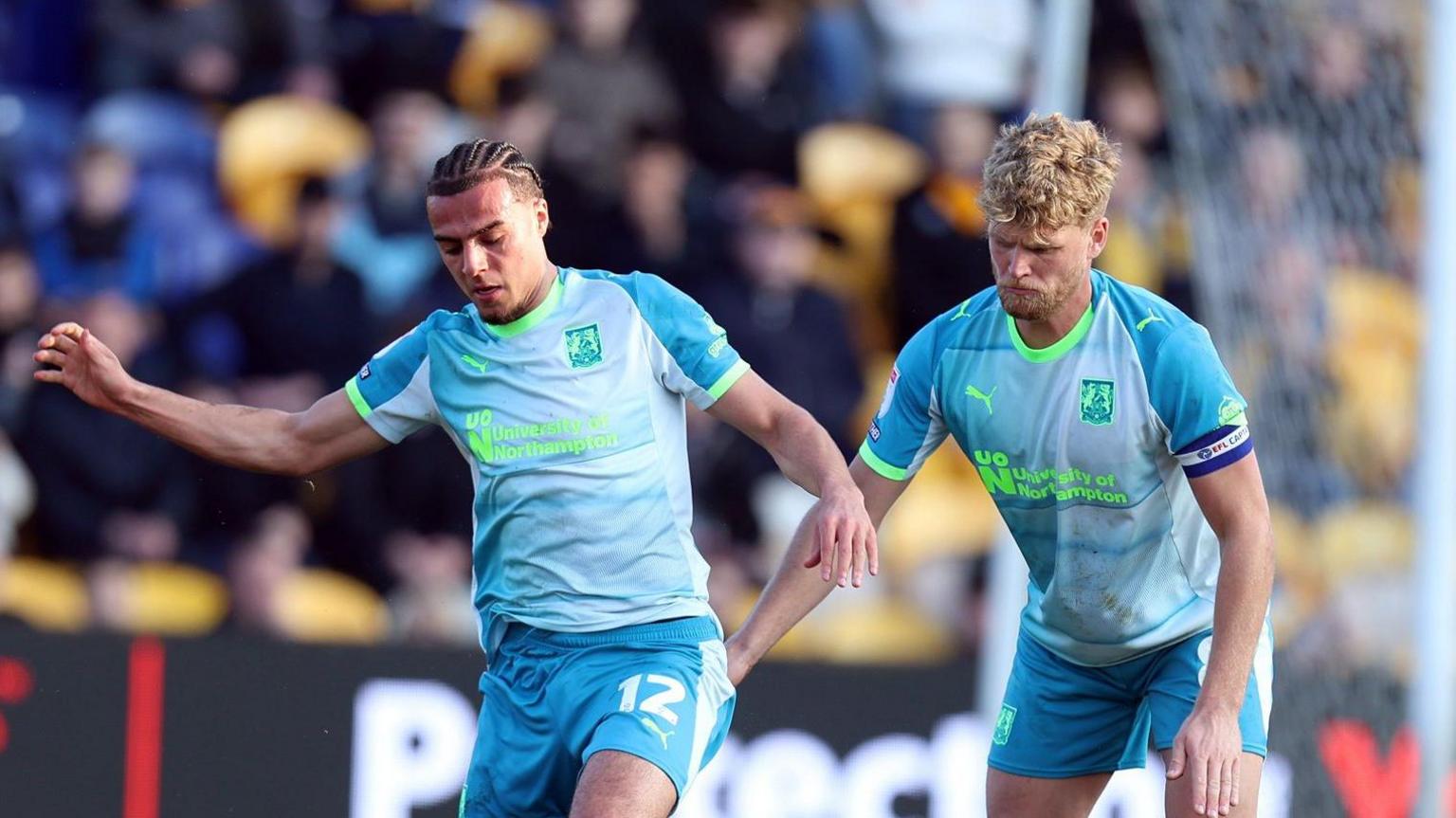 A photo of Cameron McGeehan and Nesta Guinness-Walker playing football in their light blue and fluorescent green Northampton Town third kit in March