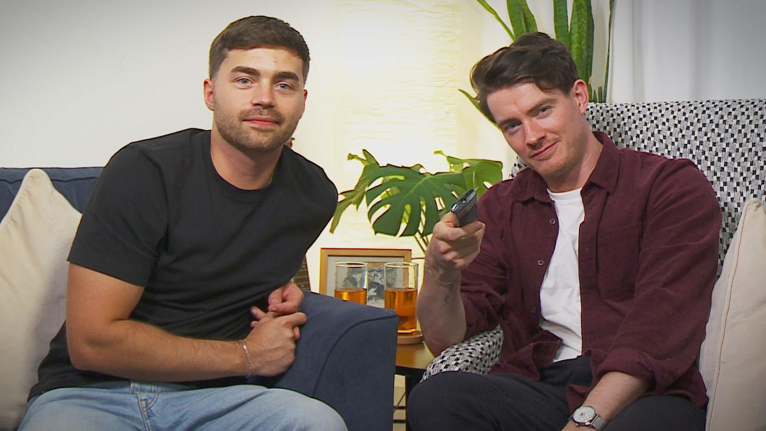 Jake and Calum sitting on armchairs, one holding a TV remote, behind them is a table with a photograph, small cheeseplant and two glasses of Irn Bru