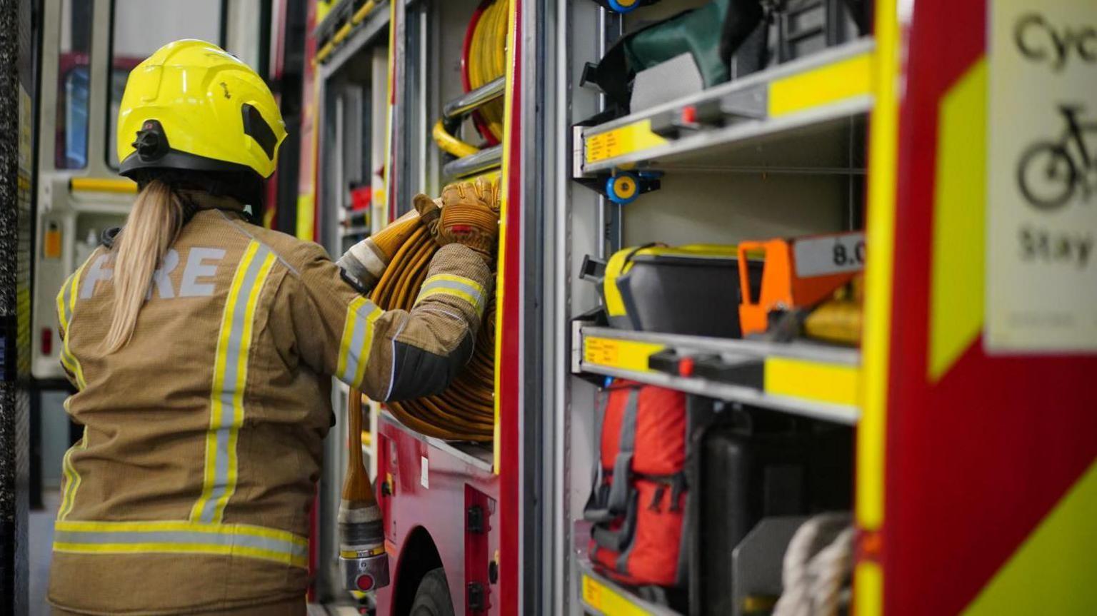 A firefighter taking a hose out of the back of a fire engine. She is wearing a yellow helmet and firefighter's hi-vis uniform