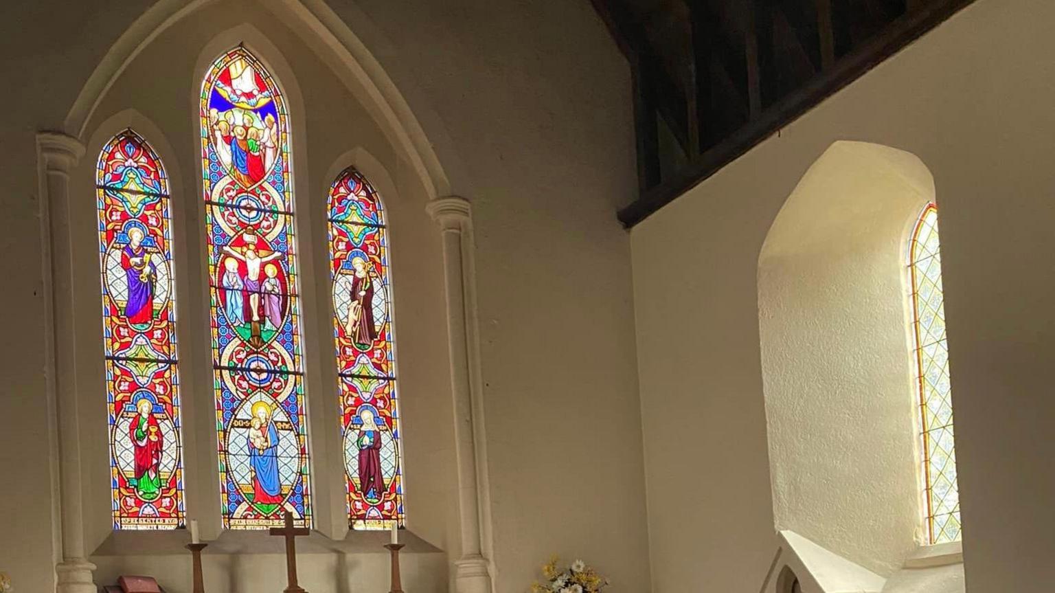 The interior of a church with an arched, stain glass window with a cross and two candles beneath it. 