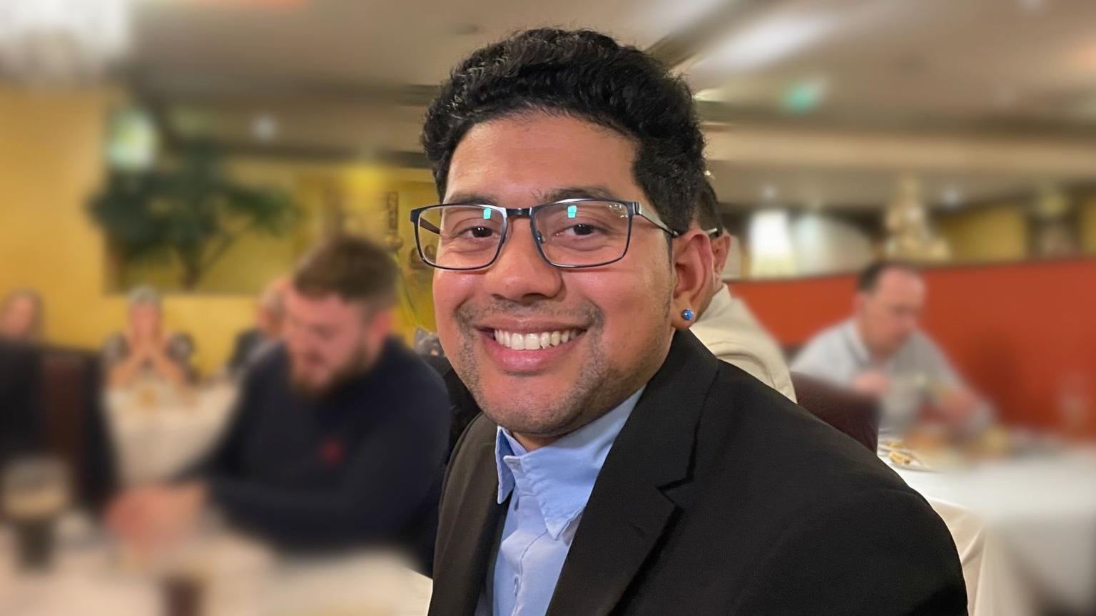 Deeshen Ruttun looking direct to camera and smiling. He has black hair and dark eyes and is wearing black-framed glasses, a black jacket and a blue shirt. Other diners in a restaurant can be seen out of focus behind him.