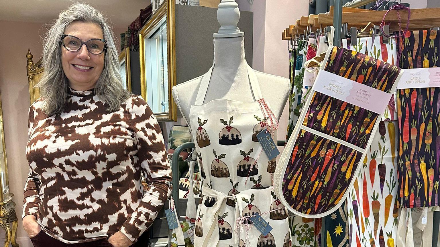 A woman wearing a brown and white patterned top, with shoulder-length grey hair and glasses, stands smiling in her shop next to a mannequin with a Christmas pudding patterned apron and some oven gloves.