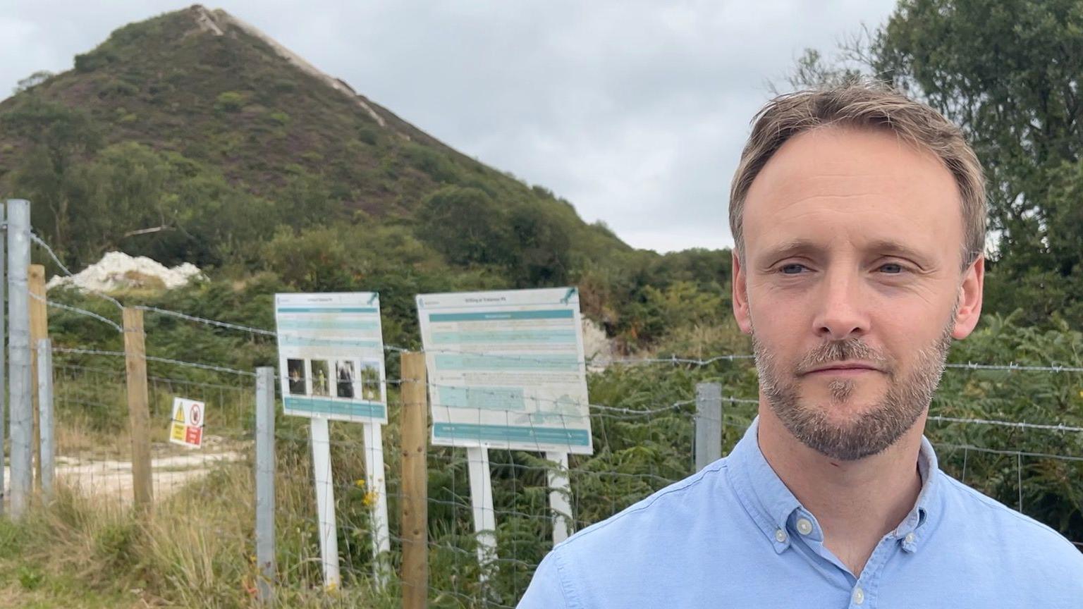A man stands in front of the waste tip known as "pointy" he wears a blue shirt and has a short beard.
