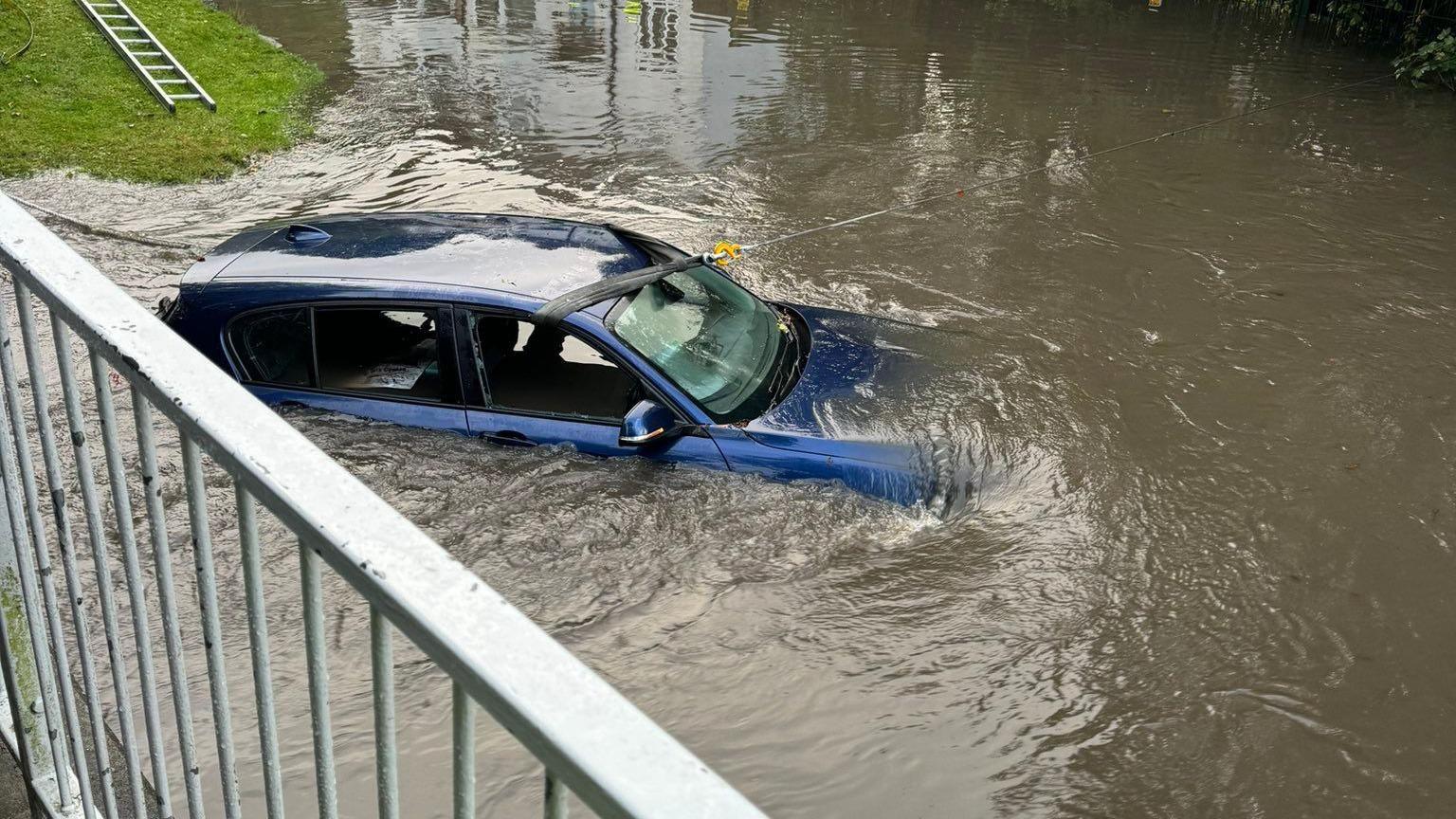 A blue car submerged in water up to the bonnet, with railings above the water