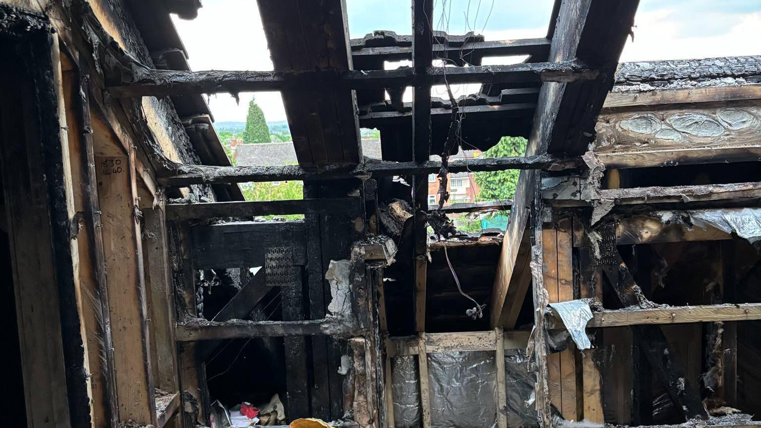 The gutted family home. All of the beams are charred and their is black everywhere. Burned items can be seen on the floor.