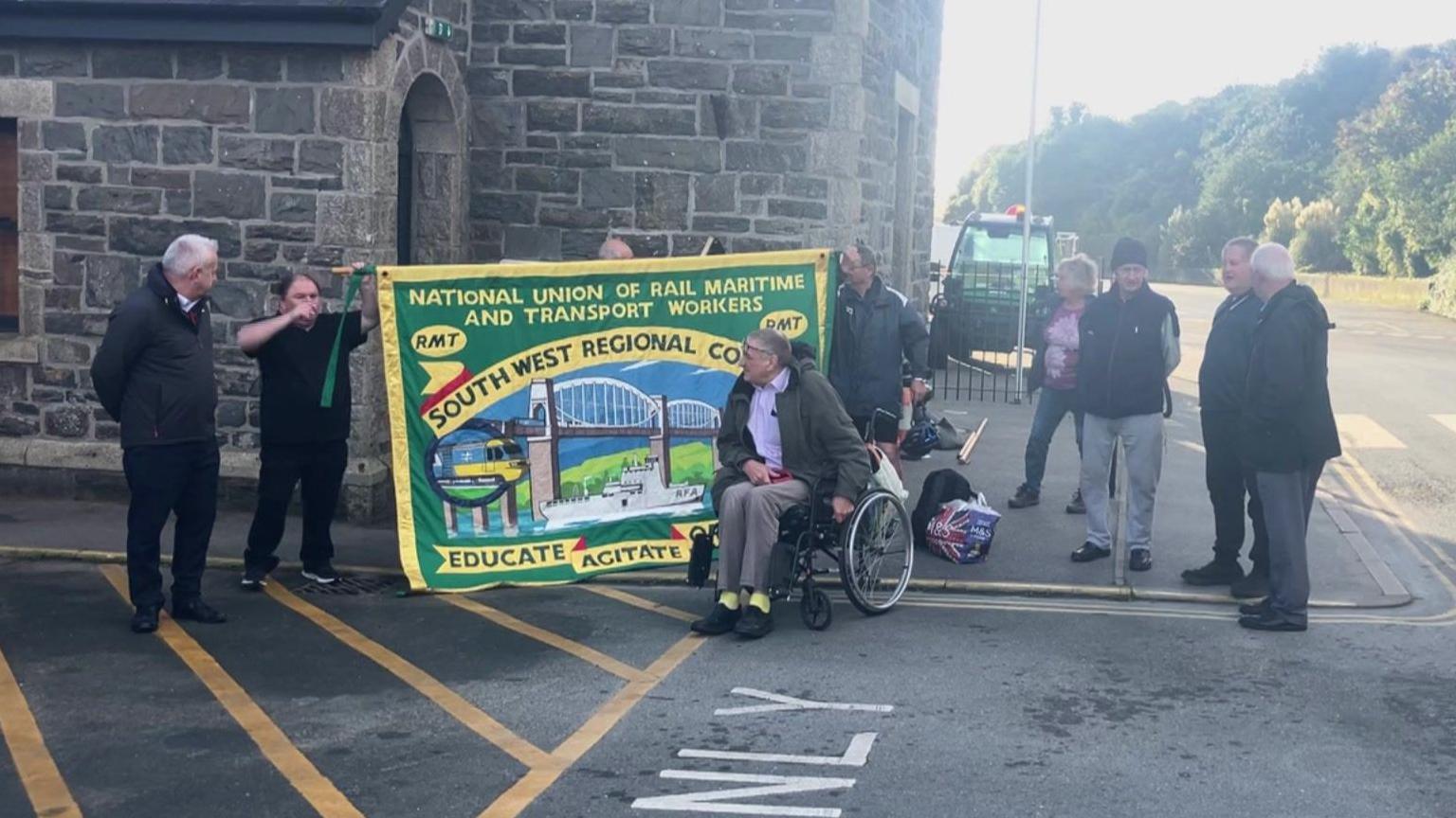 A group of nine protesters stood in front of a building next to a bus stop hold up an green and yellow RMT union banner. 