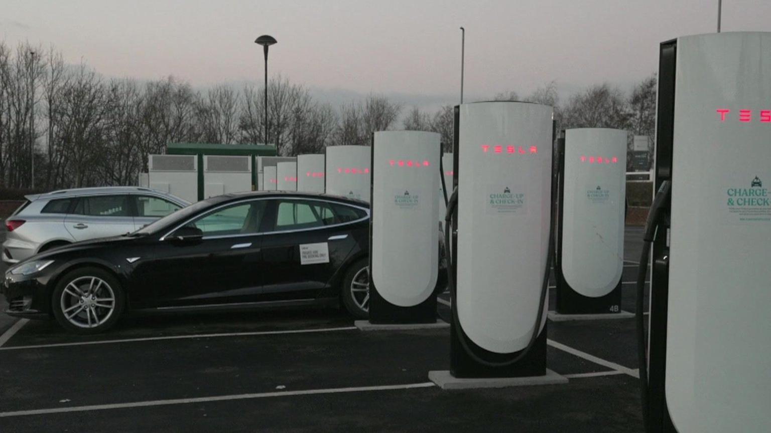 A row of charging bays in a car park, with white towers installed in every parking space. A black electric car and a white electric car are both parked up and charging.  