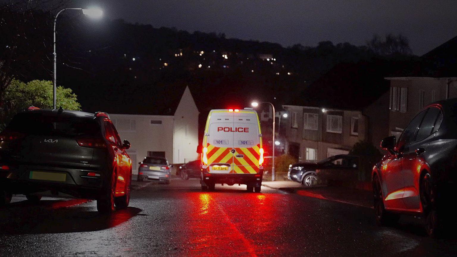 The rear of a police van, with red lights glowing off a wet road surface middle of shot, flanked by cars and houses on each side.