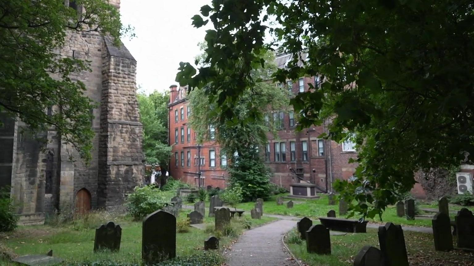 View of a wooded churchyard, gravestones surround a path, with the lower part of a stone church to the left. A large brick building backs onto the churchyard, where the light is shadowed and dim.