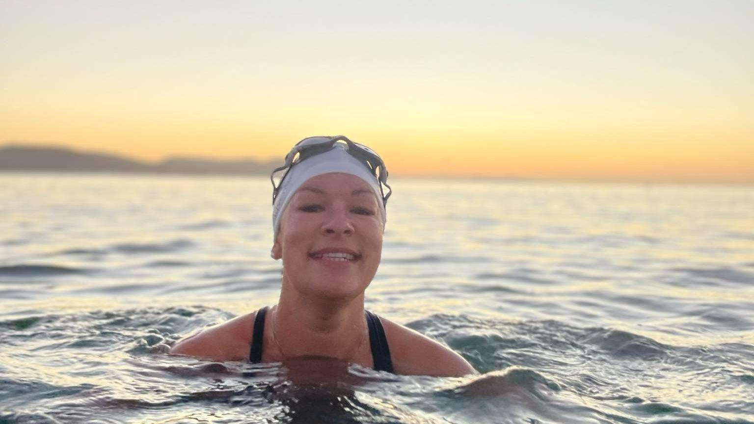 A woman in the sea smiles towards the camera against a sun rise. 